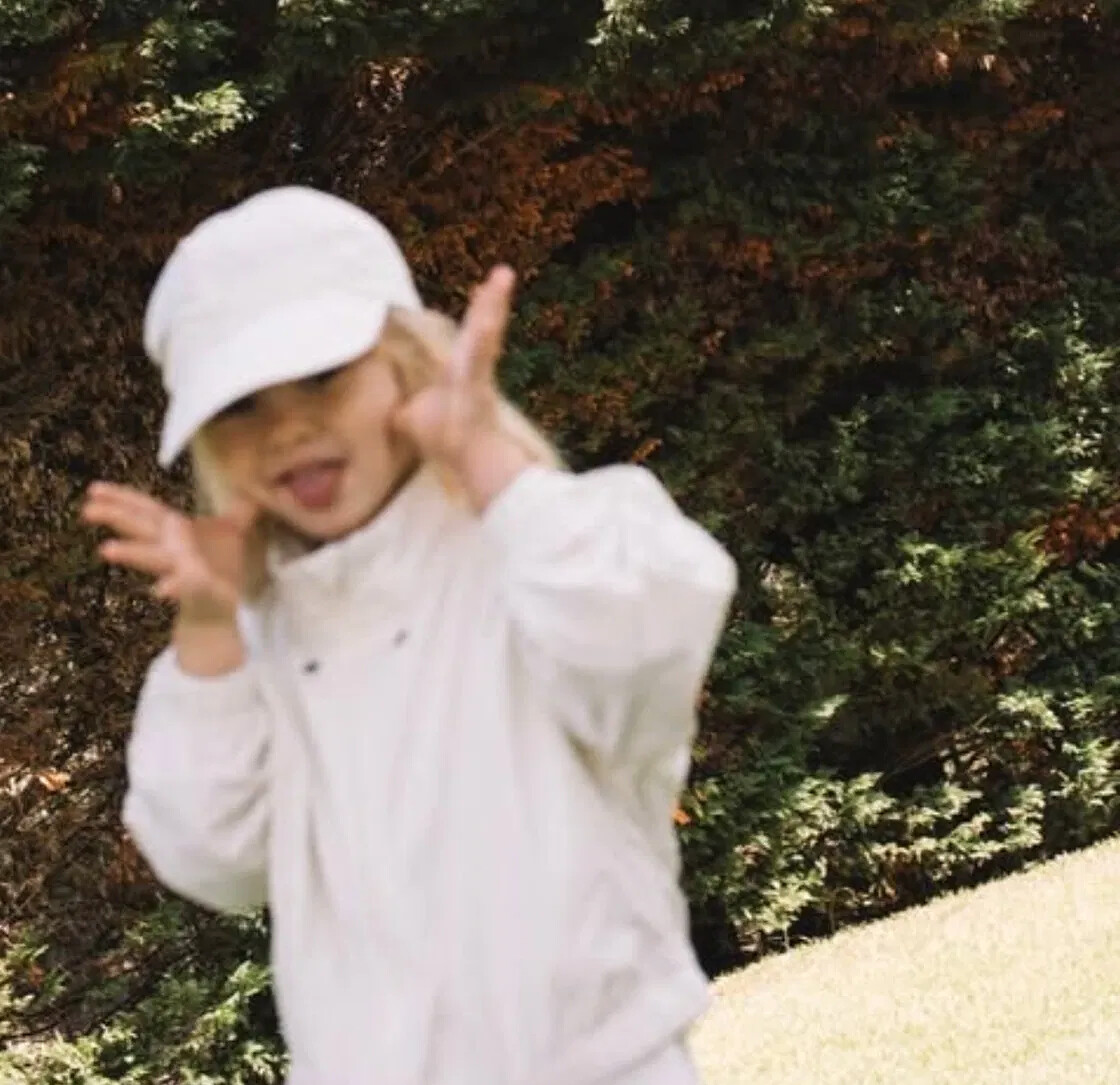 A small child in a cream bucket hat and organic-cotton pullover, mid-play against a dark hedge.