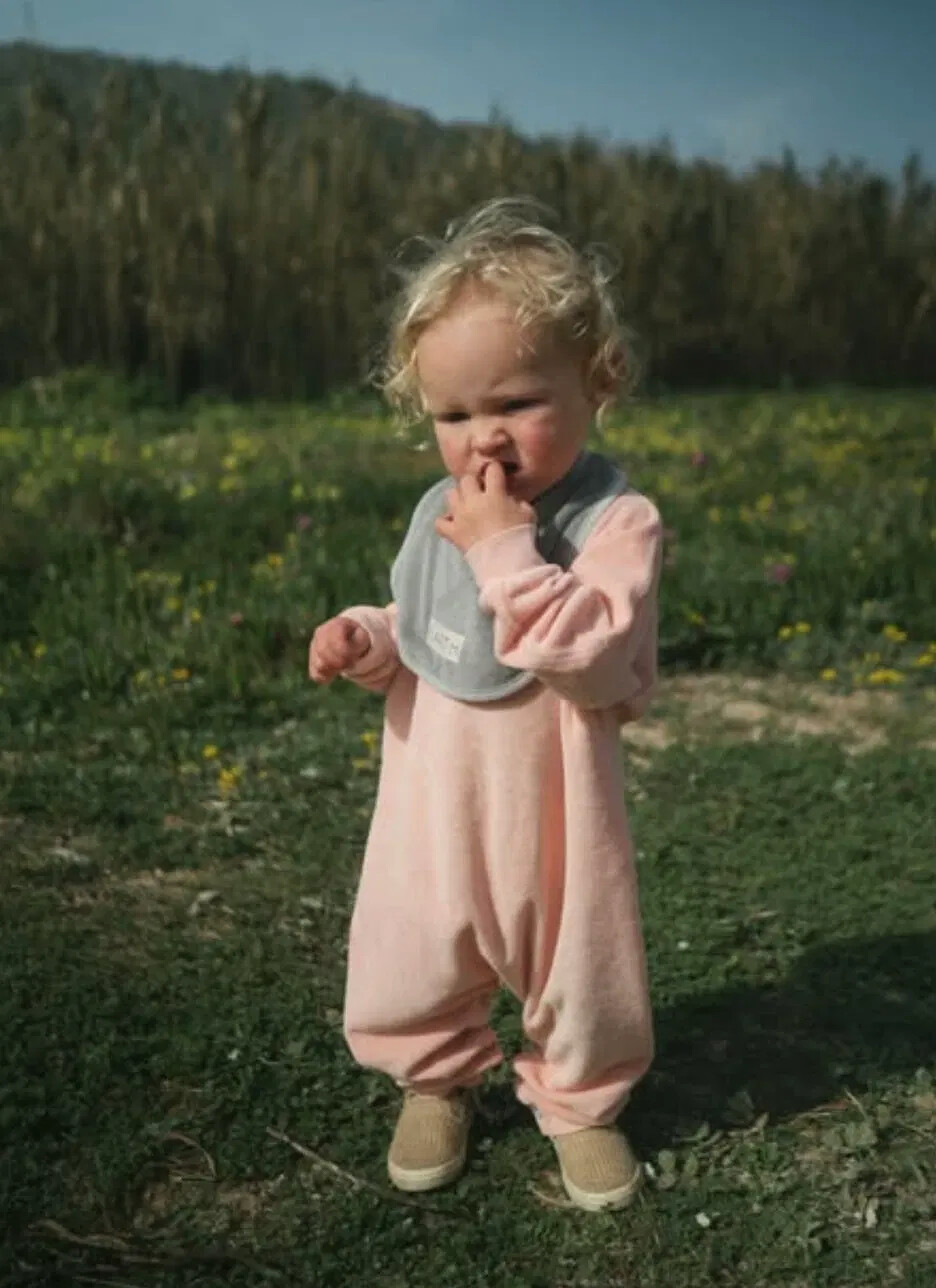 A toddler in a soft pink botanical-dyed romper and bib stands in a wildflower meadow.