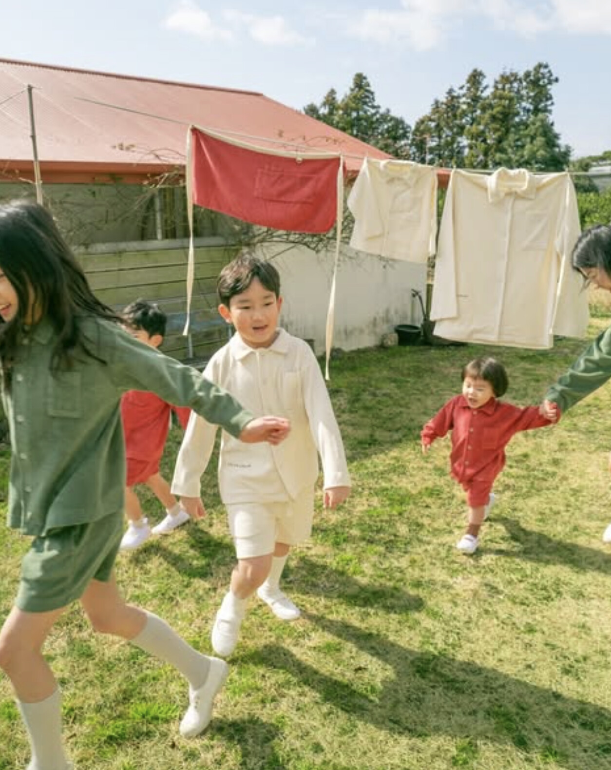 Children in olive, cream and rust play in a sunlit yard with laundry drying on the line behind them.