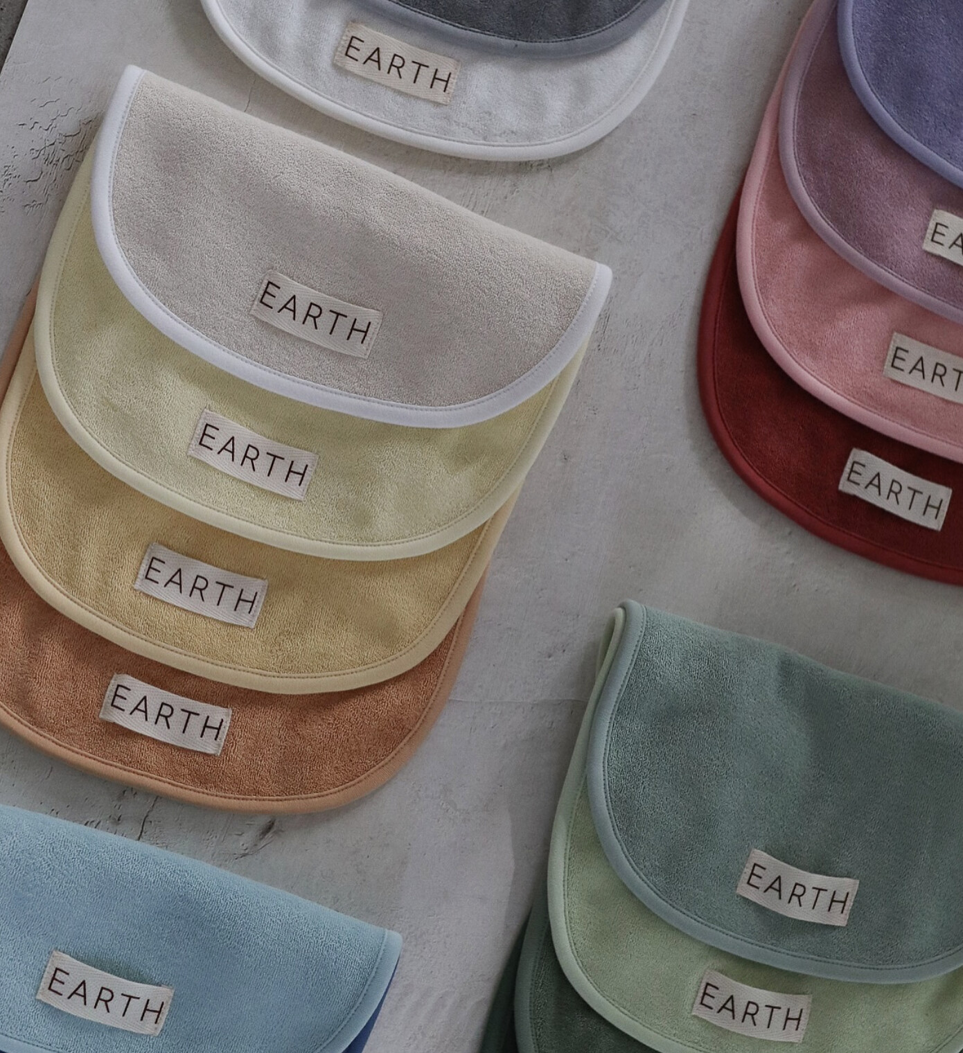 A flat-lay of terry bibs in two dozen botanical shades — sage, clay, ochre, dusty rose, slate — each labelled EARTH.