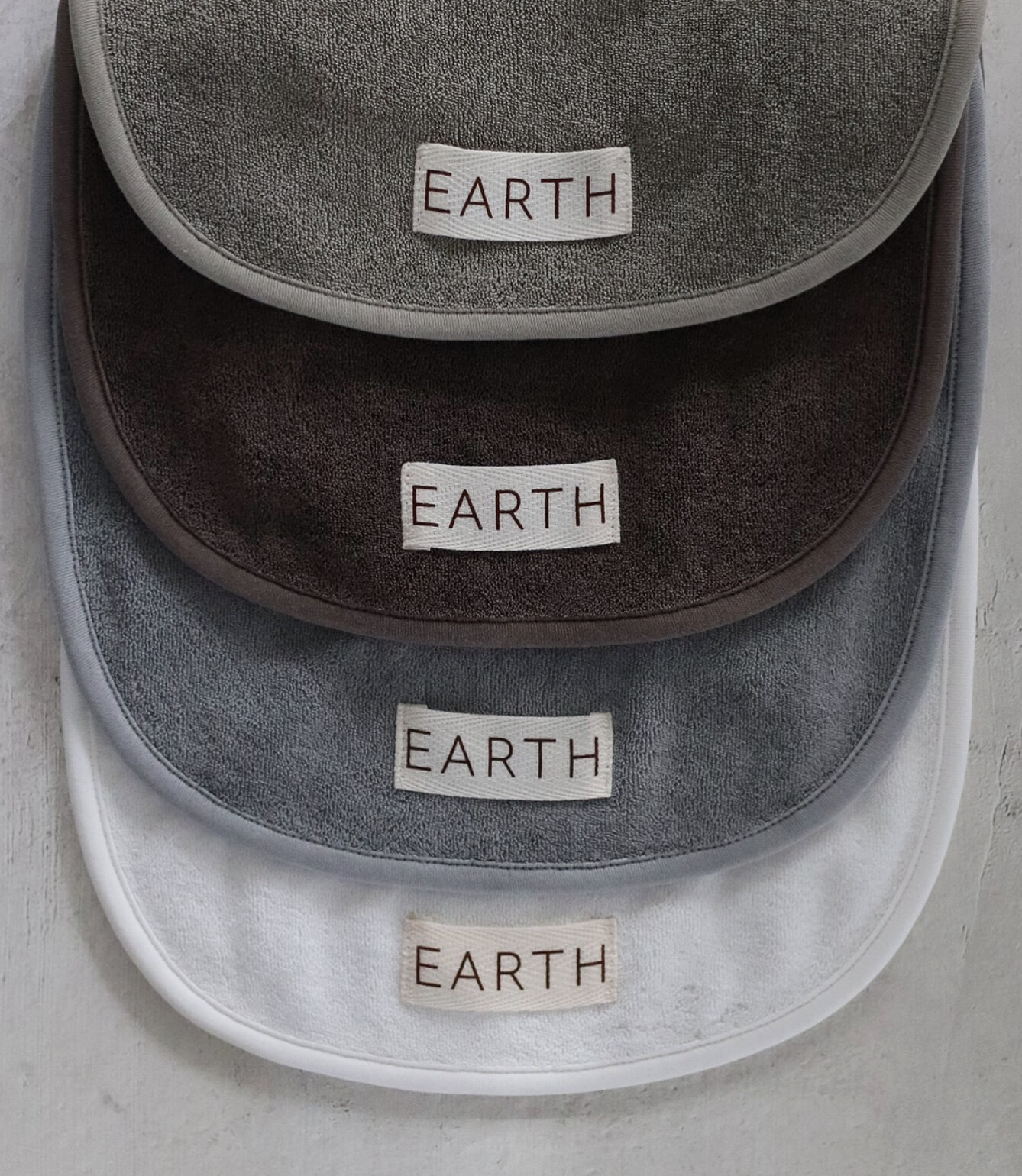 Four terry bibs stacked and fanned — sage grey, deep brown, slate blue and white — each labelled EARTH.