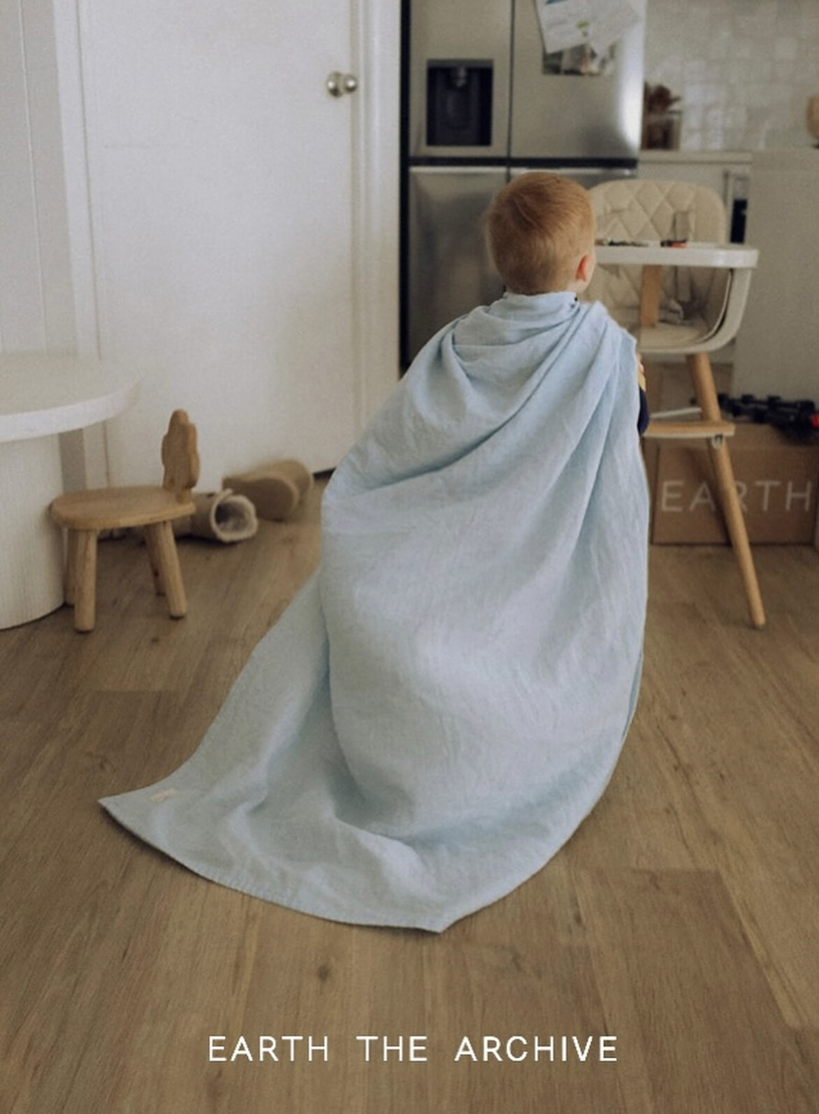 A small boy trails a pale-blue muslin blanket like a cape across a sunlit kitchen, an EARTH box beside the high chair.