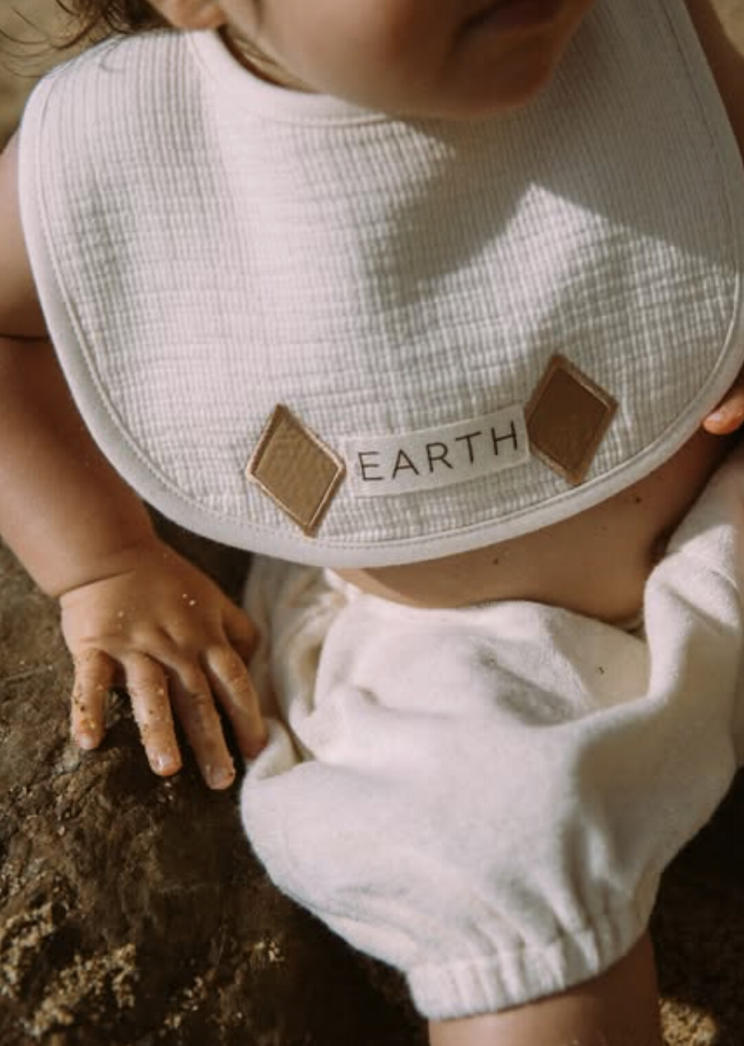 Close-up of a baby in a waffle-knit EARTH bib with twin suede diamond patches, sitting in warm sand.